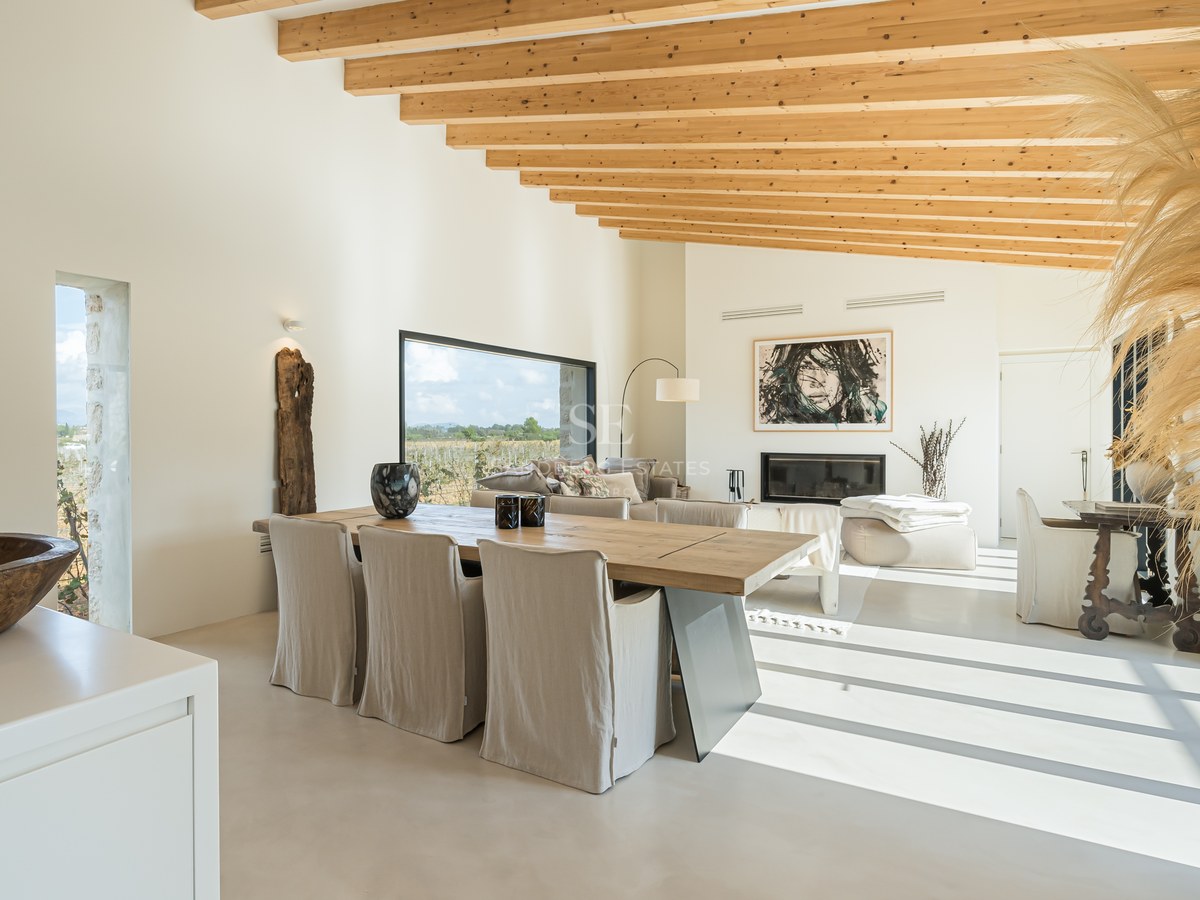 Salle à manger lumineuse avec poutres en bois, grande table en chêne et baies vitrées donnant sur la campagne.