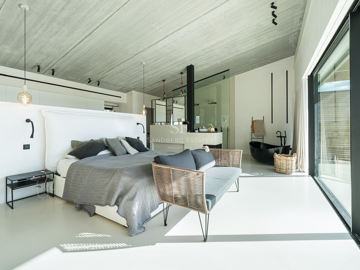 Chambre principale moderne avec lit gris, plafond en béton et baignoire îlot noire intégrée.