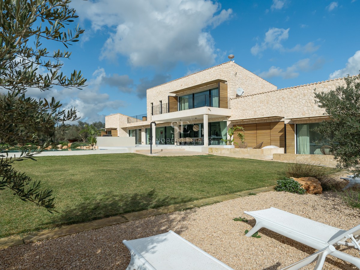 Villa contemporaine de deux étages avec murs en pierre, grandes baies vitrées et jardin sous un ciel bleu.