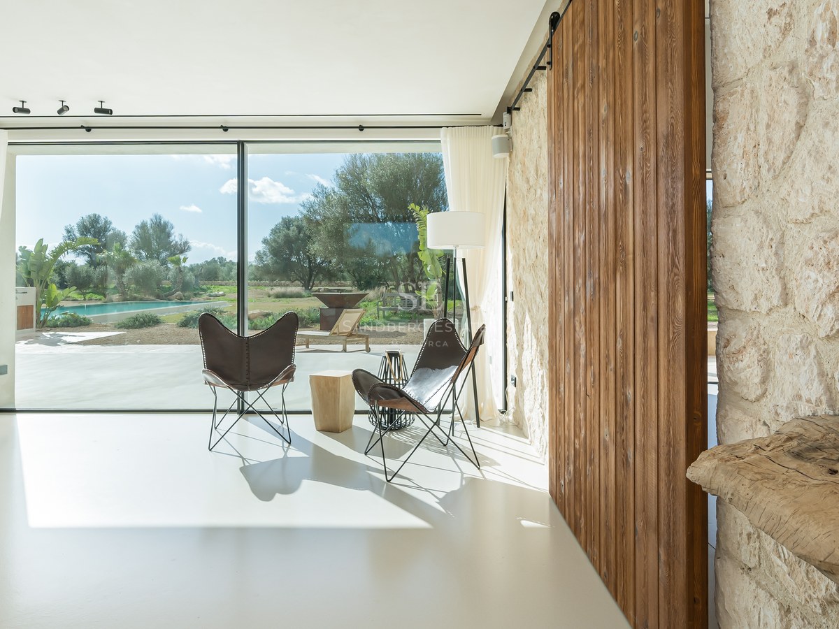 Salon lumineux avec baies vitrées, fauteuils butterfly, porte coulissante en bois et mur en pierre naturelle.