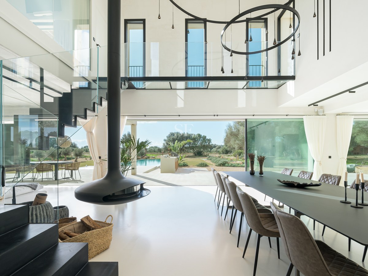 Salle à manger lumineuse à double hauteur avec cheminée suspendue noire, parois vitrées et vue sur le jardin et la piscine.