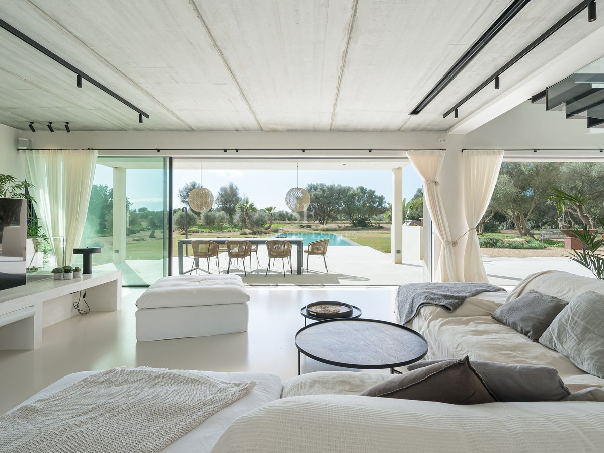 Intérieur de salon lumineux avec canapés blancs, ouvrant sur une terrasse, un coin repas extérieur et une piscine.