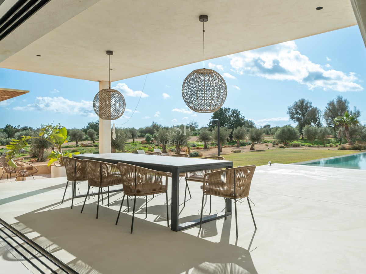 Espace repas extérieur couvert avec table moderne et chaises en rotin surplombant un jardin d'oliviers.