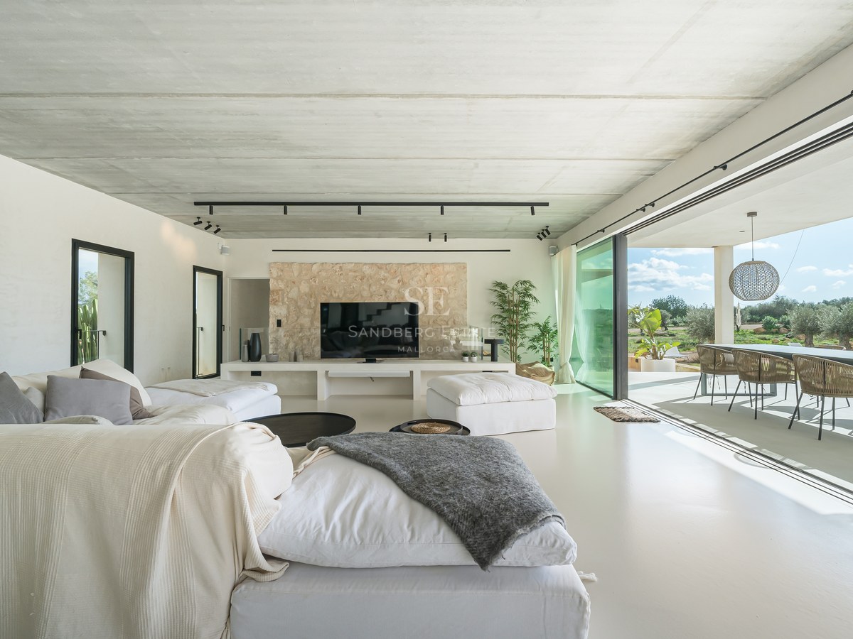 Salon moderne avec canapés blancs, plafond en béton, mur en pierre et baies vitrées ouvrant sur une terrasse.