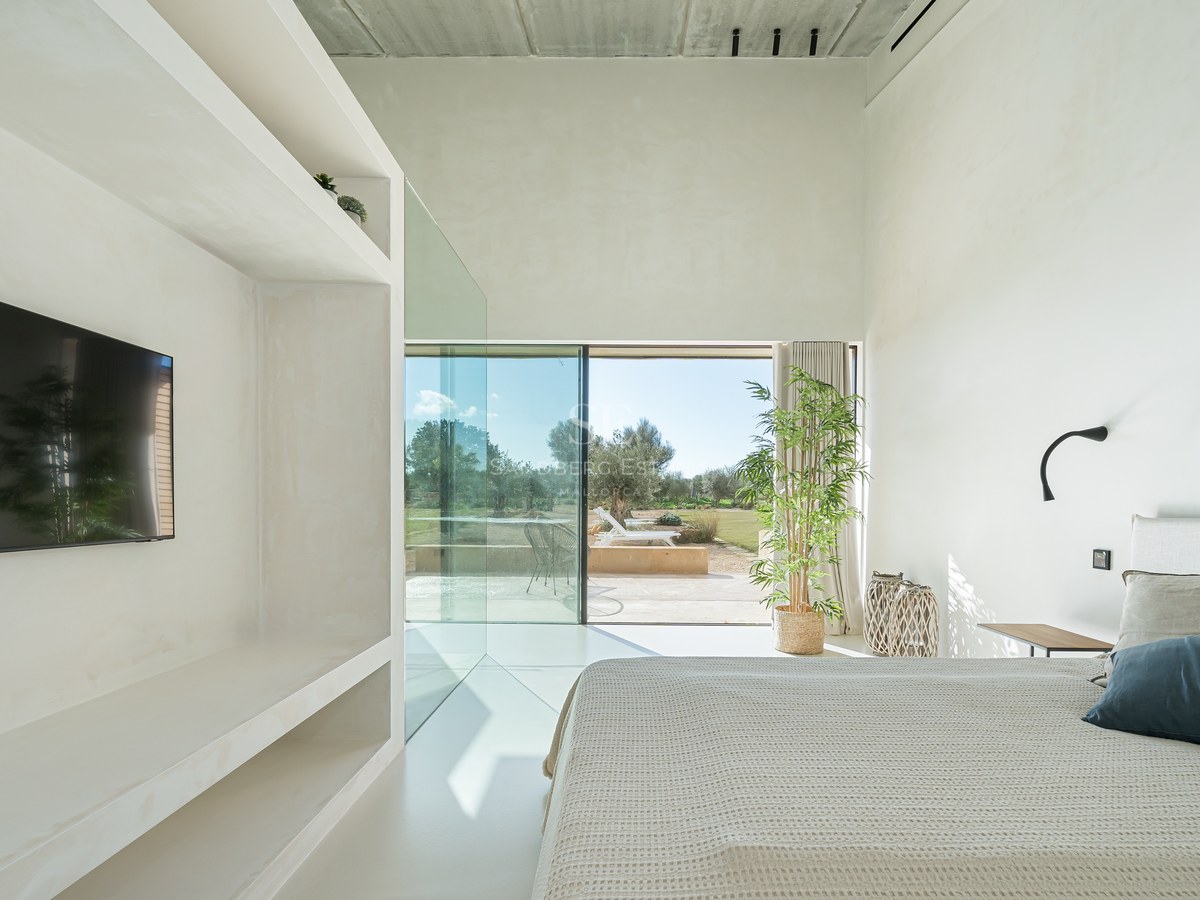 Chambre blanche minimaliste avec de grandes baies vitrées ouvrant sur une terrasse ensoleillée et un jardin.