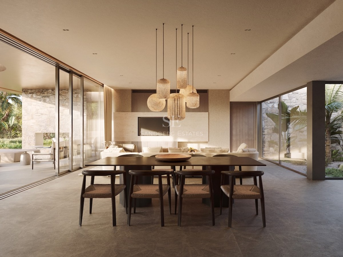 Salle à manger moderne avec table en bois sombre et suspensions en rotin, s'ouvrant sur une terrasse et un jardin.