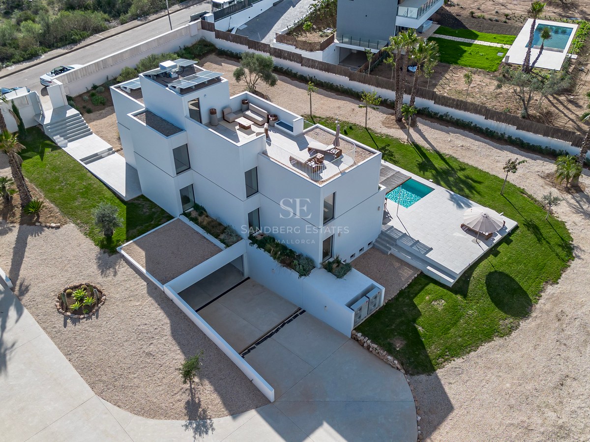 Flygfoto över en vit modern villa i tre plan med pool, anlagd trädgård och solpaneler.