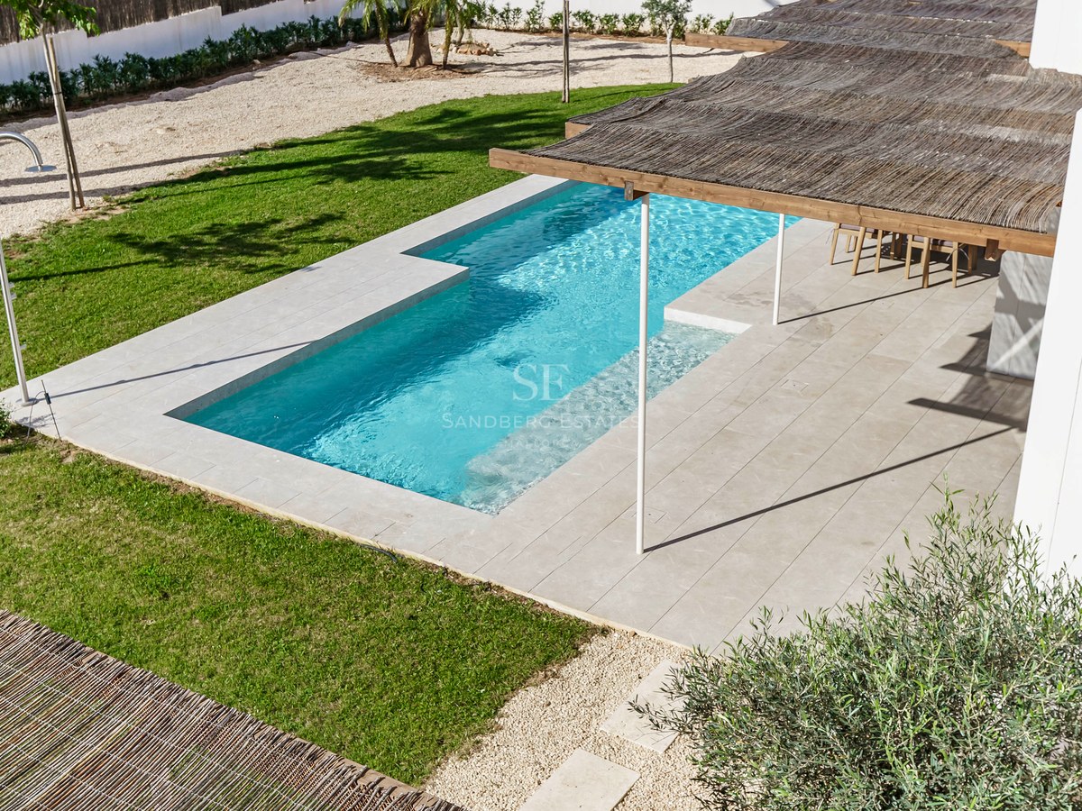 Flygvy över en turkos rektangulär pool omgiven av stenterrass, gräsmatta och en pergola med vasstak.