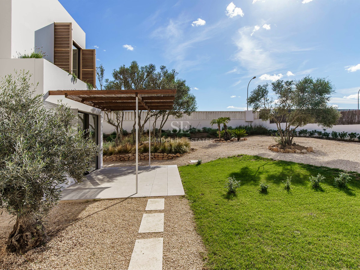 Exteriör av modern villa med olivträdgård, gräsmatta, grusgångar och träpergola under en klarblå himmel.