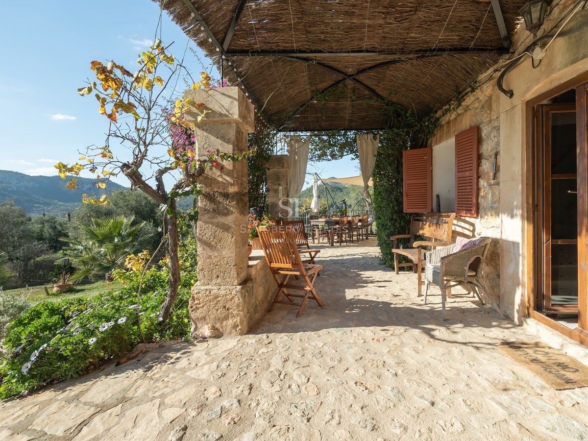 Terrasse en pierre rustique avec toit de chaume, meubles en bois et vue sur les collines verdoyantes.