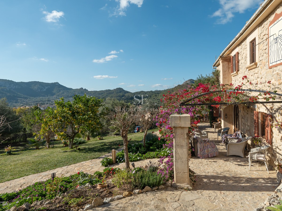 Terrasse en pierre avec mobilier extérieur, bougainvilliers roses et vue sur les montagnes sous un ciel bleu.