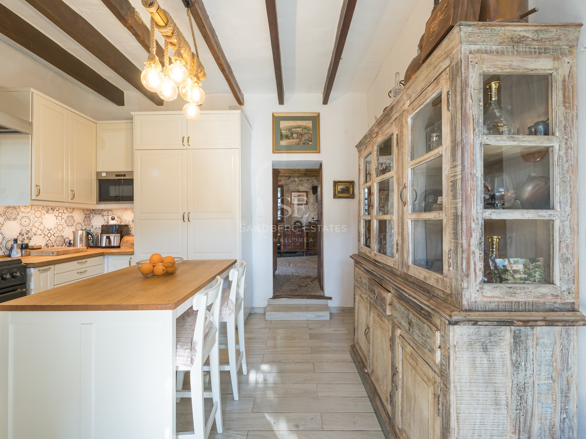 Cuisine lumineuse avec poutres de plafond en bois sombre, armoires blanches, îlot avec tabourets et grand buffet vintage.
