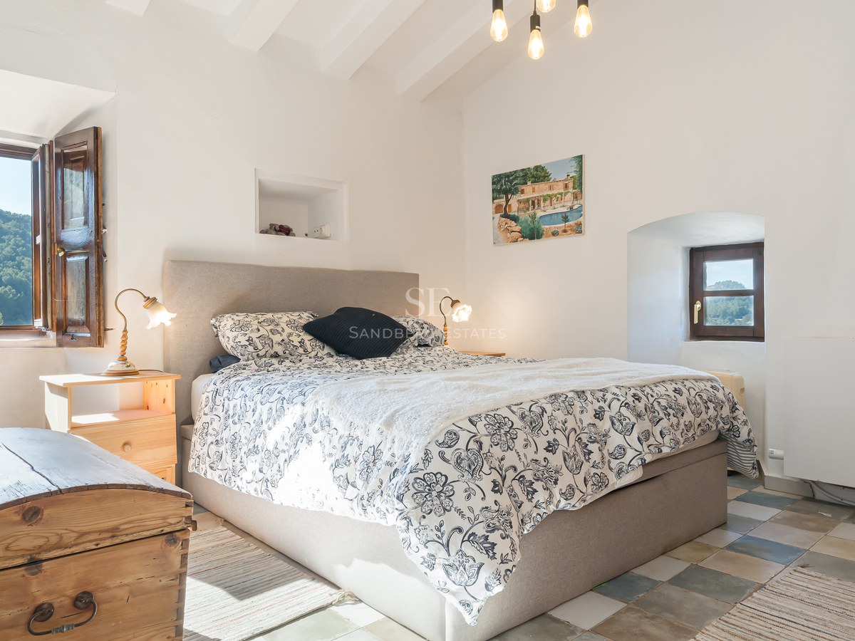 Chambre lumineuse avec poutres blanches, lit rembourré, linge de lit floral et tables de chevet en bois rustique.