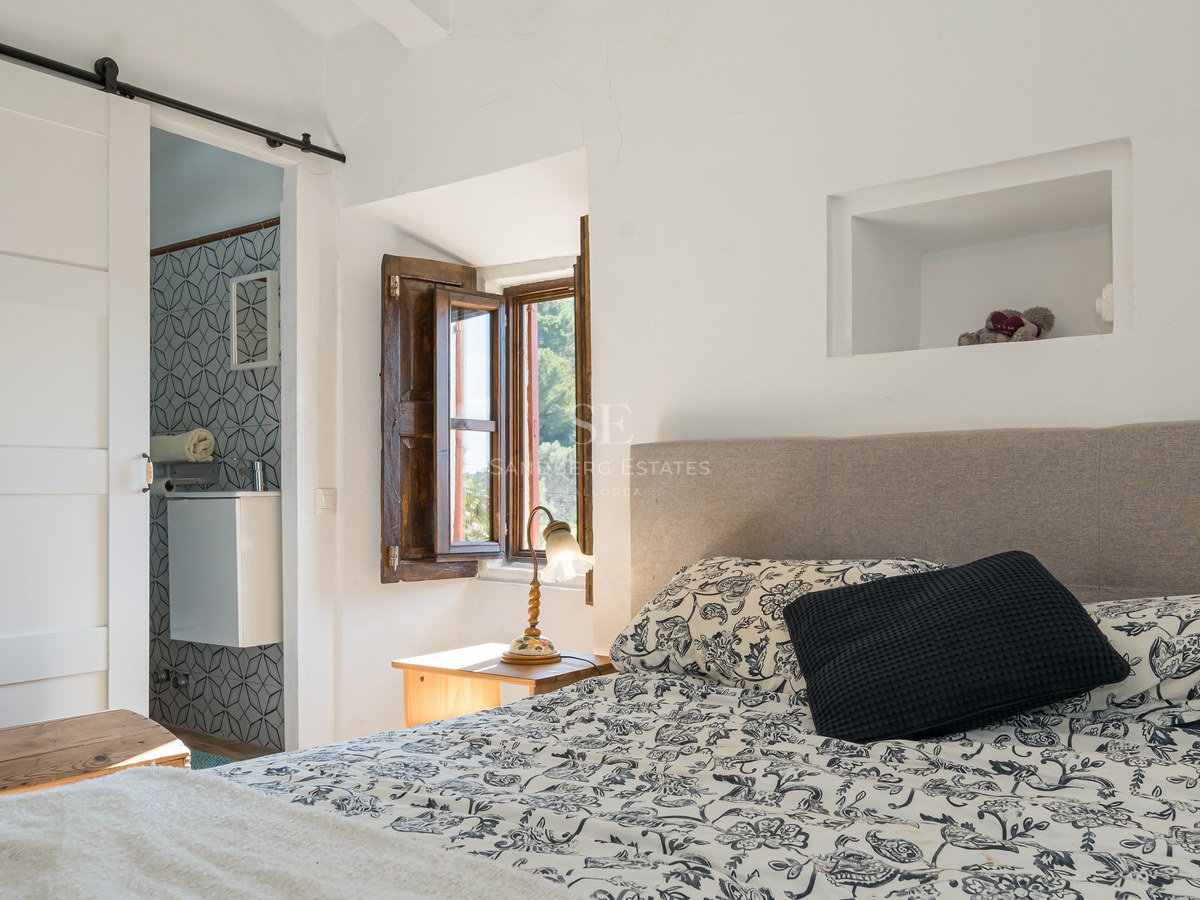 Chambre ensoleillée avec couvre-lit à motifs, volet en bois traditionnel et vue sur une salle de bain carrelée.