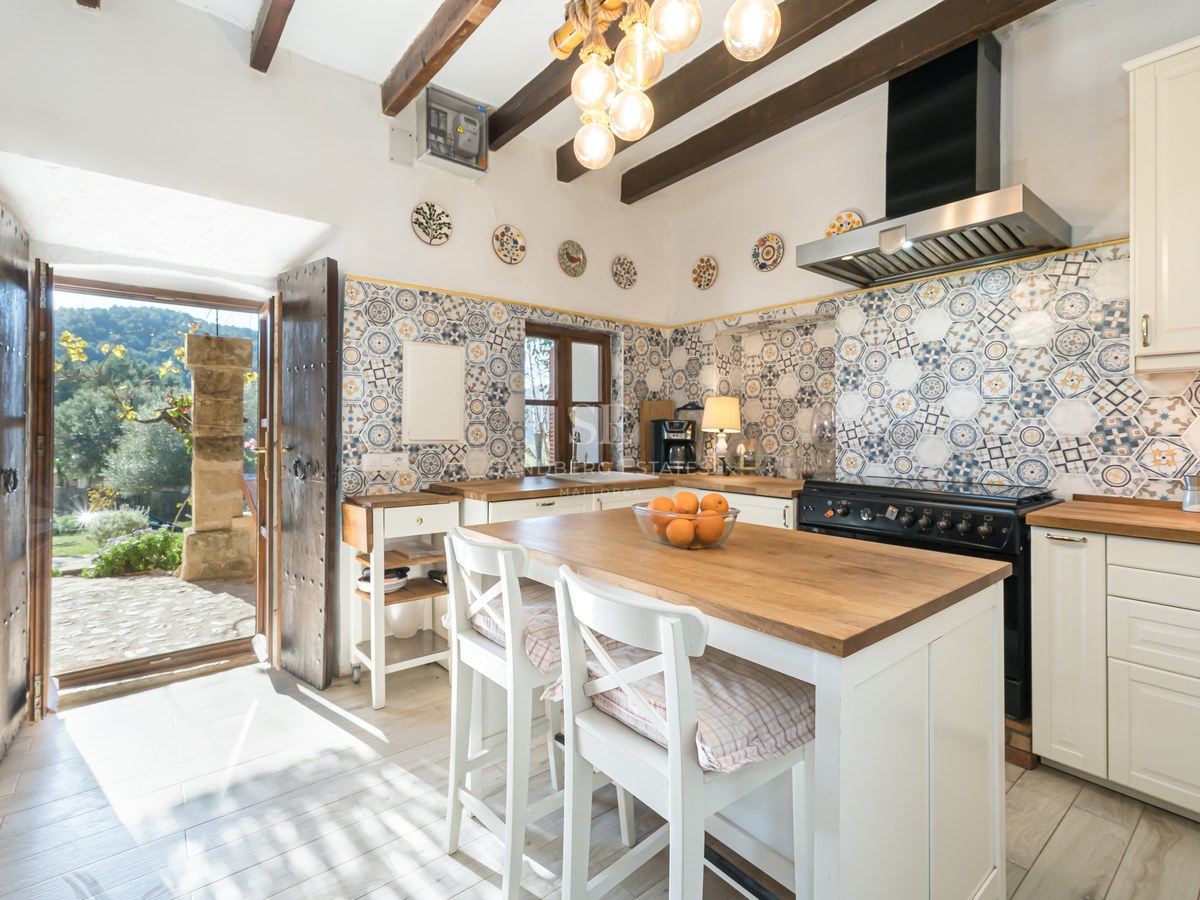 Cuisine rustique avec poutres en bois, carreaux artisanaux, îlot central avec tabourets et vue sur cour.