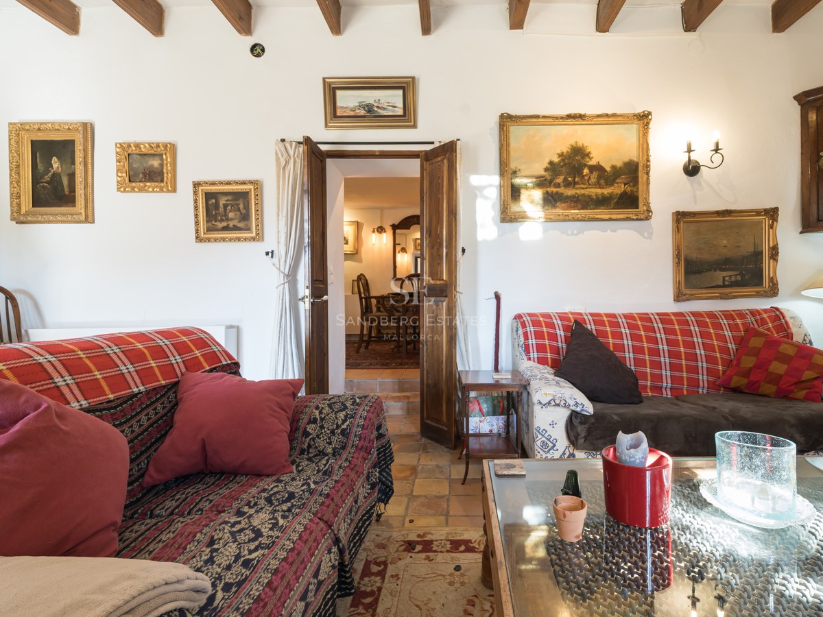 Salon de style rustique avec sol en terre cuite, poutres en bois sombre et tableaux classiques sur murs blancs.