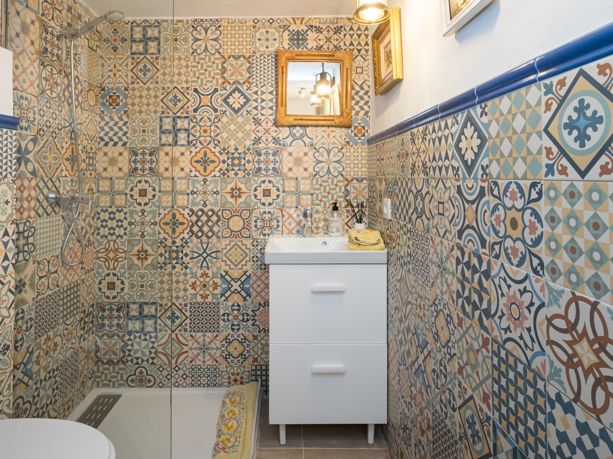 Salle de bain avec carrelage à motifs géométriques bleus et ocres, meuble vasque blanc et douche vitrée.