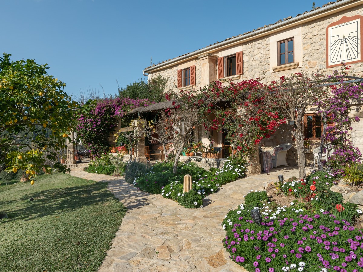 Extérieur d'une maison en pierre avec volets en bois, allée pavée et bougainvilliers en fleurs sous un ciel bleu.