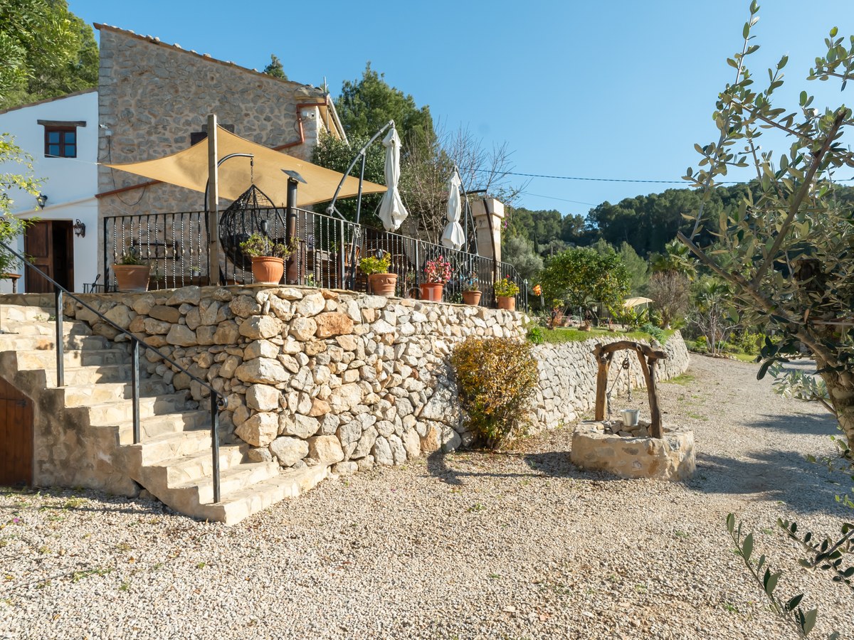 Terrasse surélevée en pierre avec voile d'ombrage, ferronnerie et puits traditionnel dans un paysage rural.