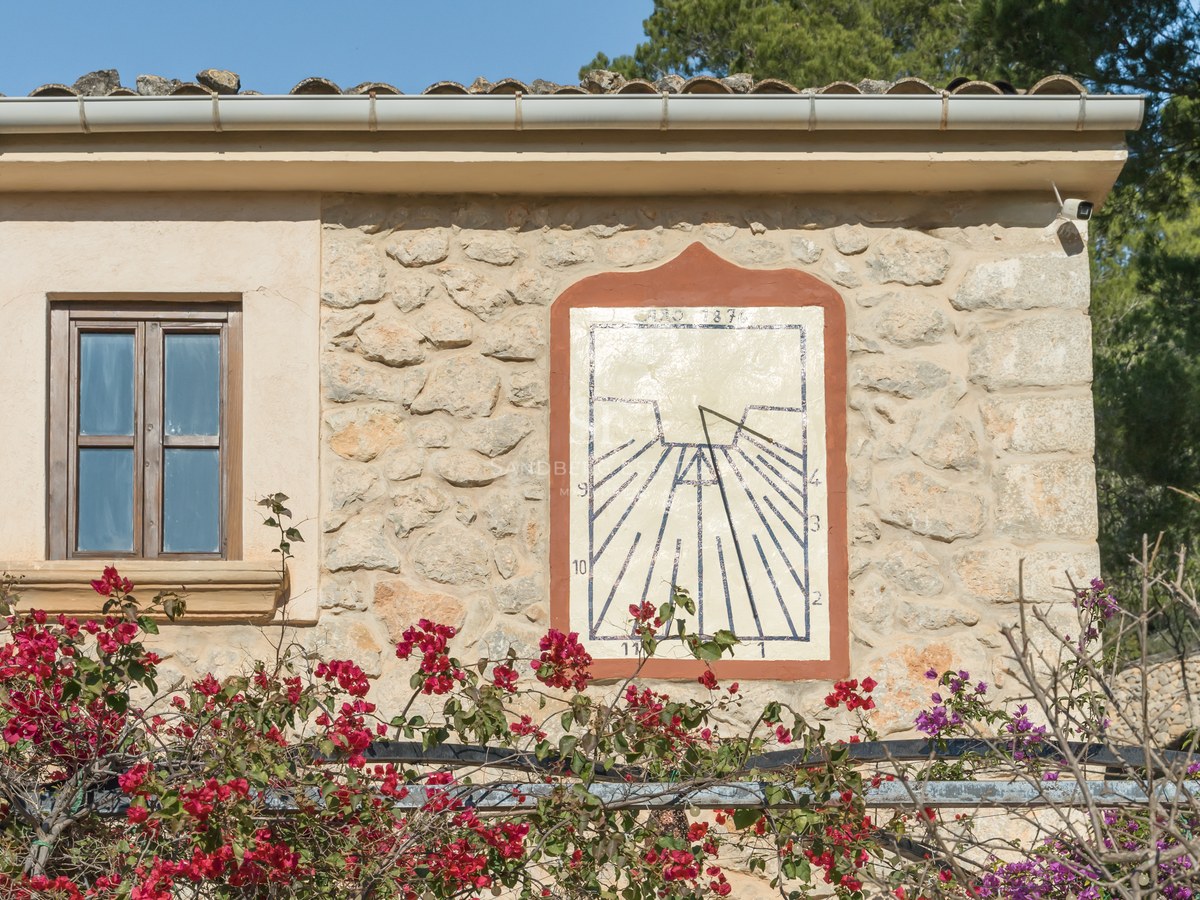 Cadran solaire artisanal sur une façade en pierre naturelle avec des bougainvilliers rouges sous le soleil.