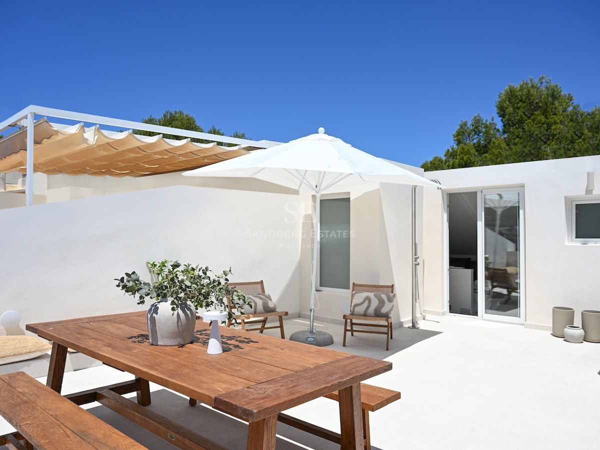 Terraza blanca soleada con mesa de comedor de madera, sombrilla y tumbonas bajo un cielo azul despejado.