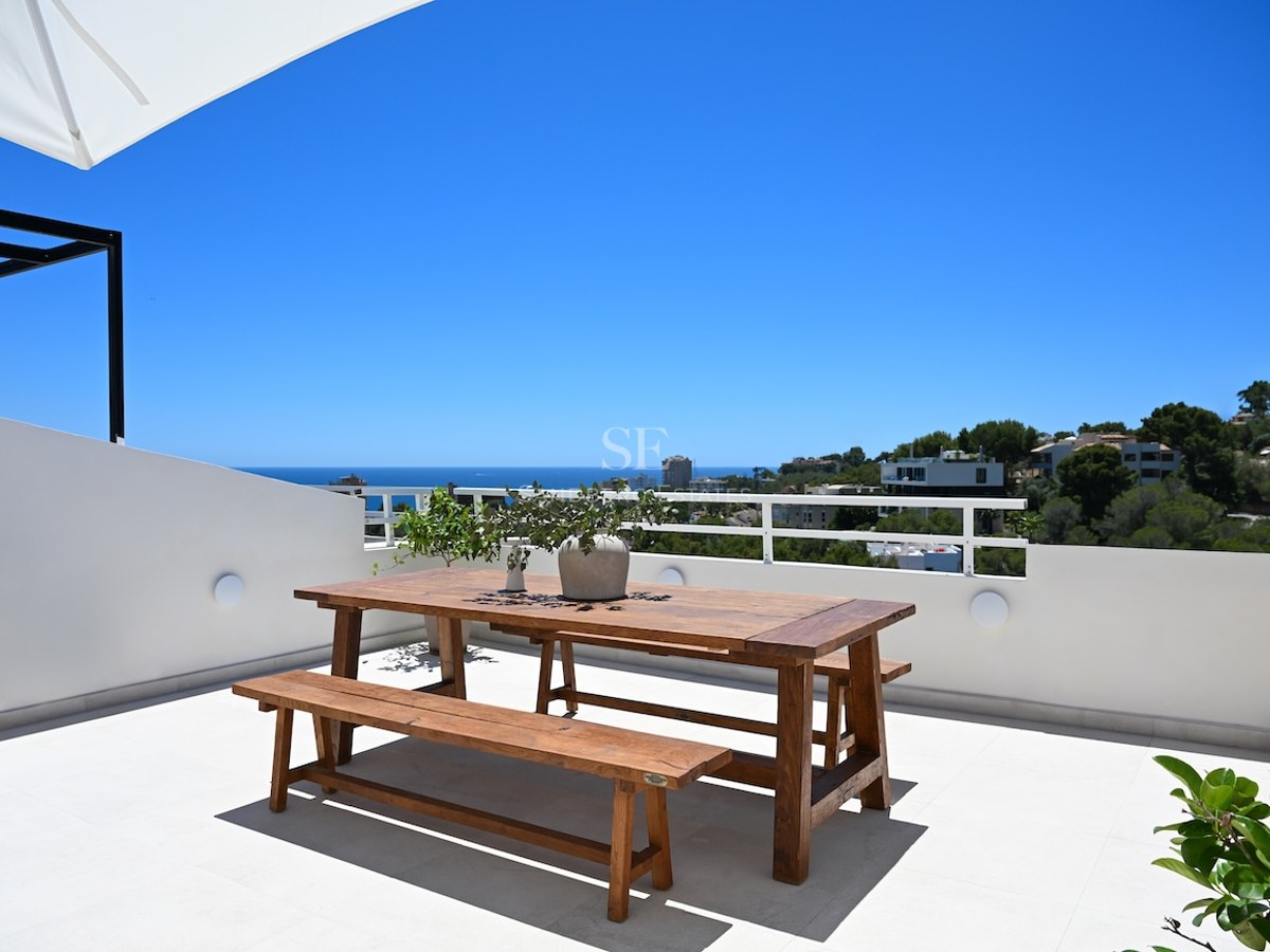 Mesa de comedor de madera en terraza blanca con vistas al mar Mediterráneo bajo un cielo azul despejado.