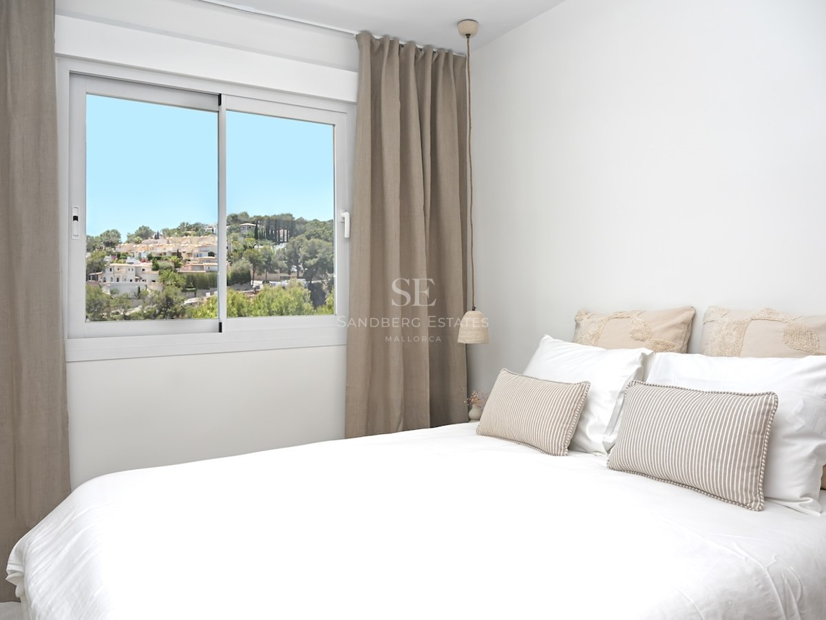 Dormitorio moderno con ropa de cama blanca, cortinas color topo y ventana con vistas a un pueblo mediterráneo.