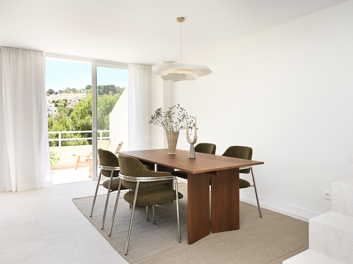 Comedor moderno con mesa de madera, sillas verde oliva y grandes ventanales con acceso a una terraza soleada.