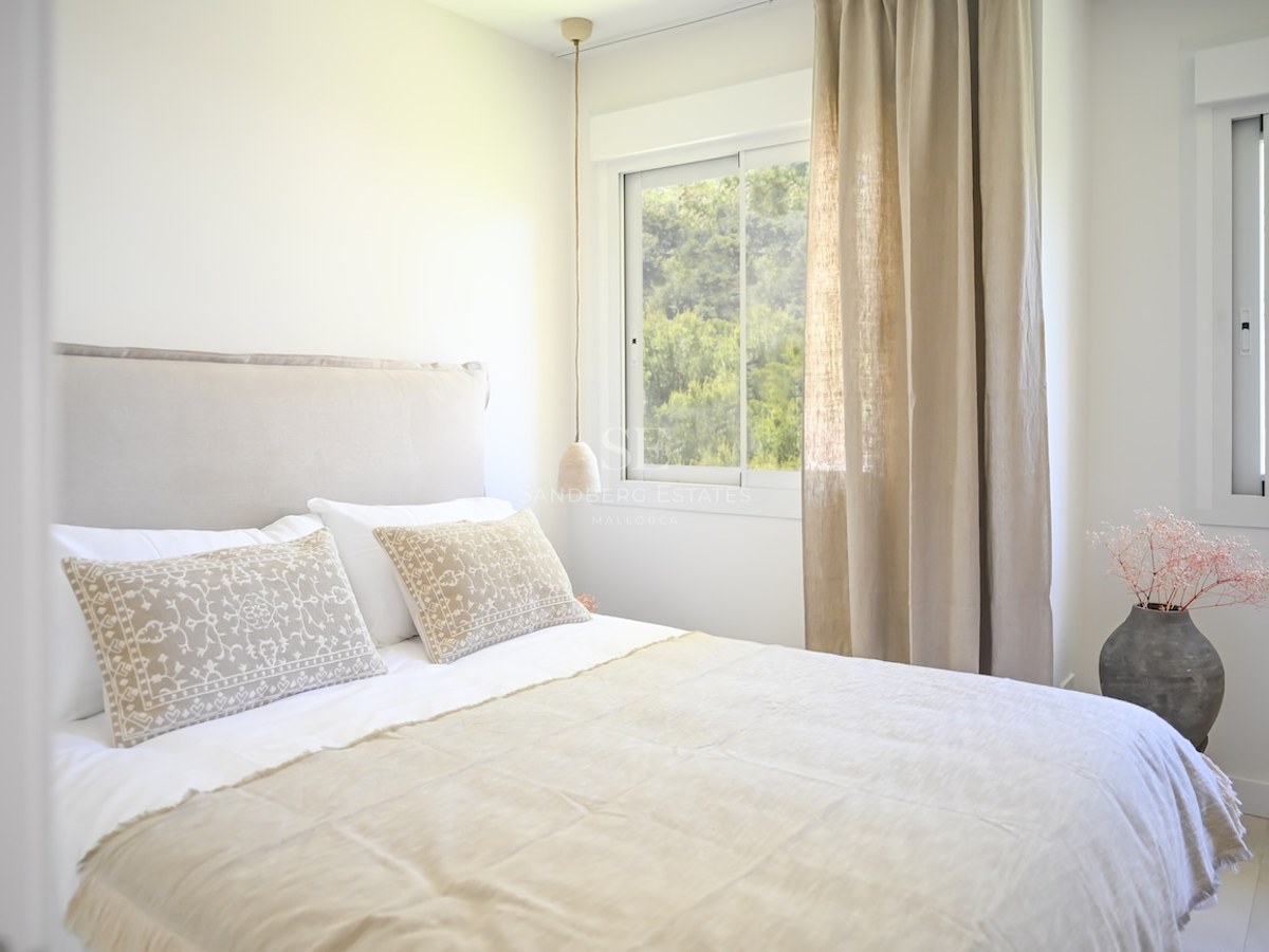 Dormitorio luminoso con cama doble, cabecero tapizado, cortinas de lino beige y vistas al jardín mediterráneo.