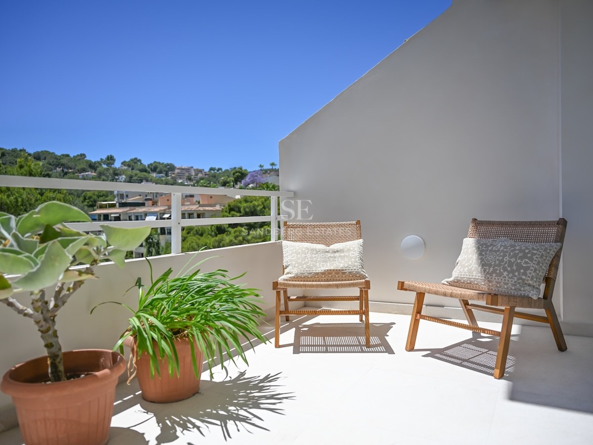 Dos sillas de mimbre en una terraza blanca y soleada con vistas a una colina mediterránea verde bajo un cielo azul.