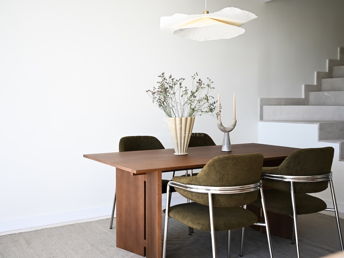 Mesa de comedor de madera con sillas verde oliva y lámpara de diseño frente a una escalera de piedra blanca.