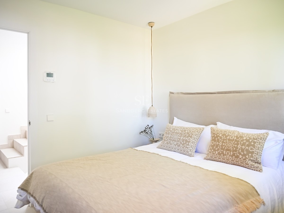 Dormitorio minimalista con textiles neutros, cabecero acolchado y lámpara colgante junto a paredes blancas.