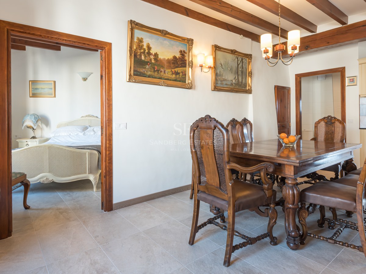 Salle à manger avec table en bois sculpté, chaises en cuir et rotin, plafond à poutres et vue sur une chambre.
