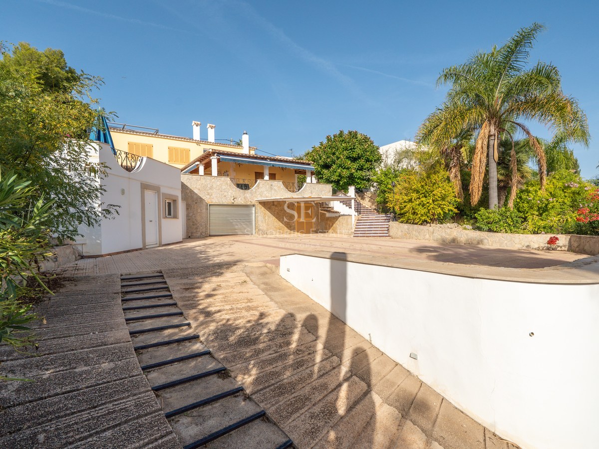 Gepflasterte Einfahrt mit Garage, umgeben von mediterraner Bepflanzung und Palmen.