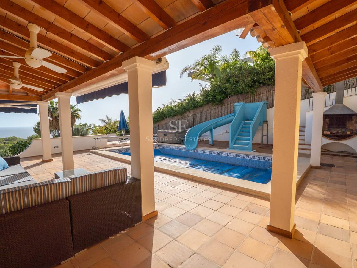 Überdachte Terrasse mit Holzbalken und Sitzgelegenheiten mit Blick auf einen blauen Pool mit Rutsche und Palmen.