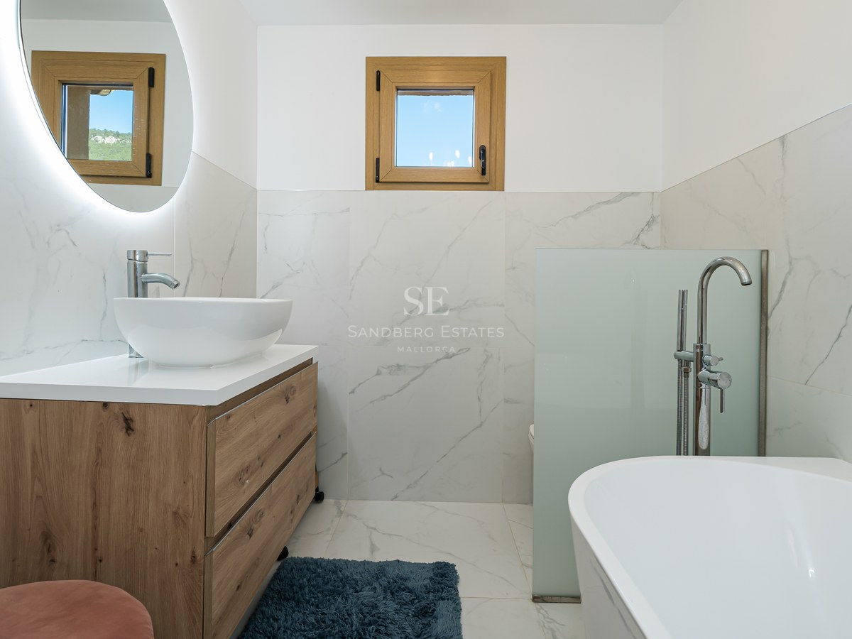 Baño moderno con paredes de mármol, mueble de madera, lavabo de sobremesa, espejo retroiluminado y bañera blanca.