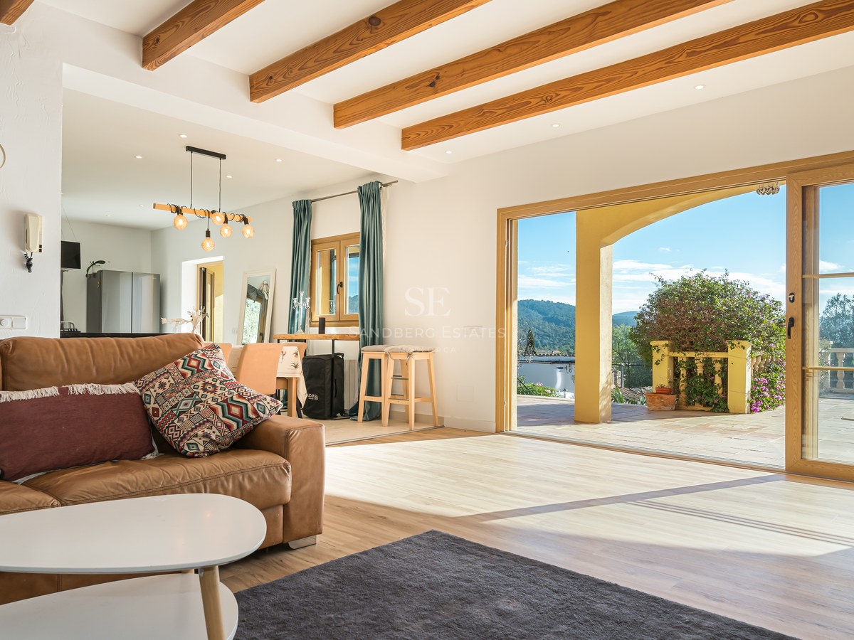 Salón con sofá de cuero marrón, vigas de madera y puertas correderas que dan a una terraza con vistas a la montaña.