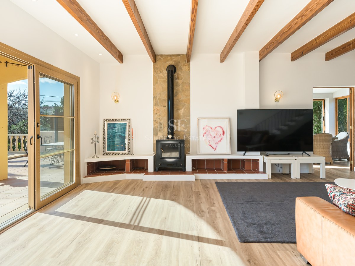Salón moderno con vigas de madera, chimenea de leña y puertas correderas que dan a una terraza soleada.