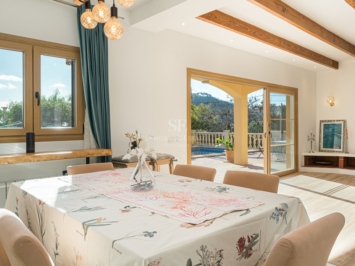 Comedor luminoso con vigas de madera, chimenea de piedra y puertas de cristal hacia una terraza con piscina.