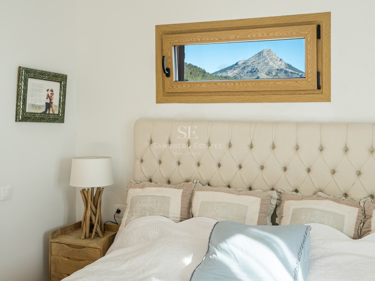 Cama con cabecero capitoné beige junto a una ventana pequeña que enmarca una montaña escarpada.