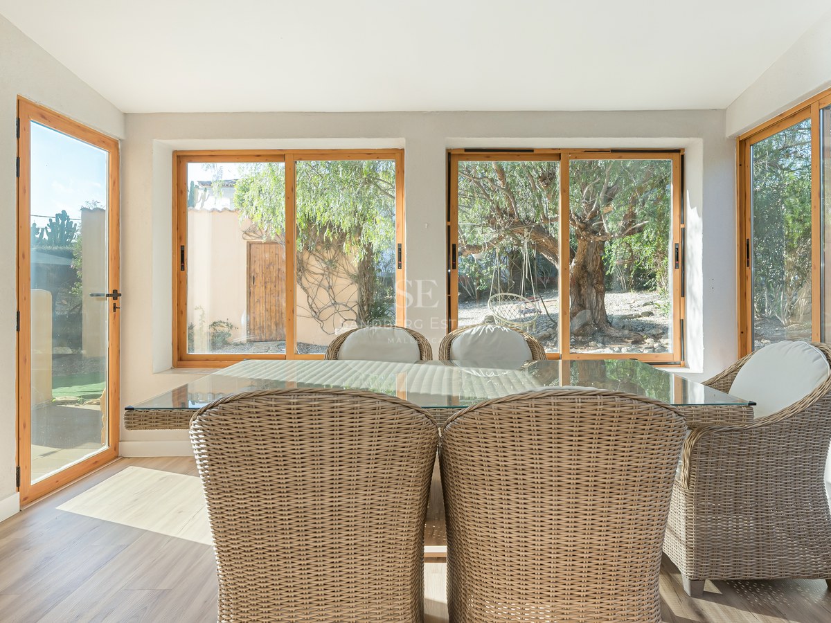 Comedor interior soleado con sillas de mimbre y mesa de cristal con vistas a un jardín con árboles mediterráneos.