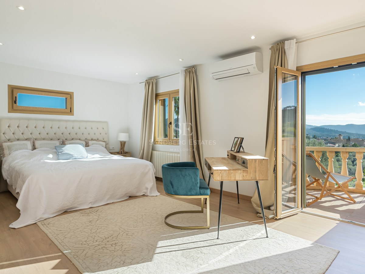 Dormitorio principal luminoso con cama grande, escritorio de madera y balcón con vistas a las montañas.