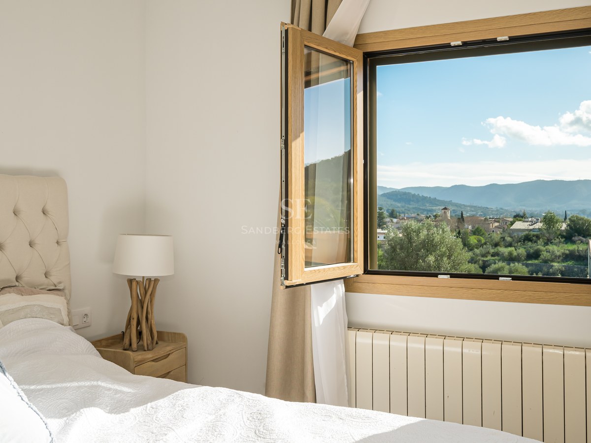 Dormitorio acogedor con cama de cabecero acolchado y ventana abierta con vistas panorámicas a un pueblo y montañas.