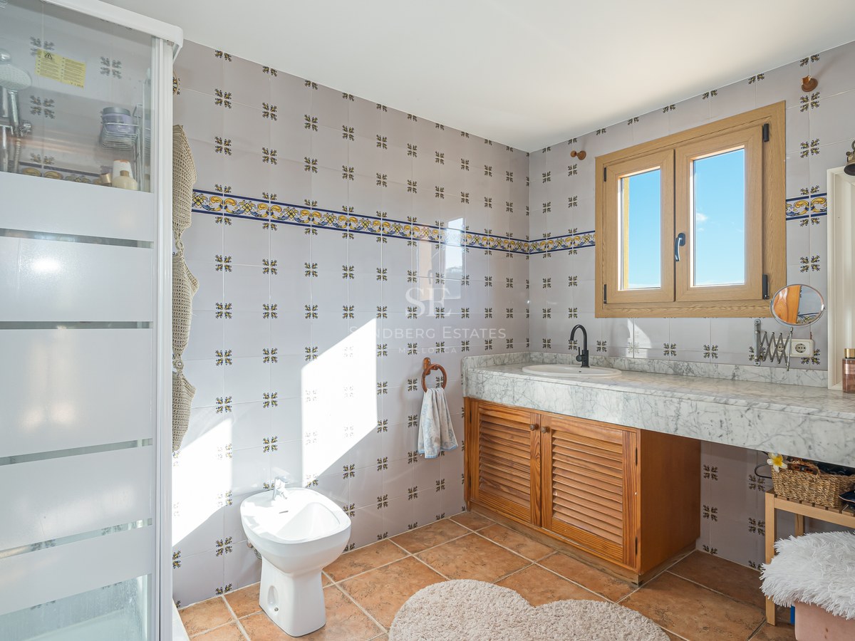 Baño con azulejos decorados, encimera de mármol blanco, mueble de madera, bidé y suelo de terracota bajo luz natural.
