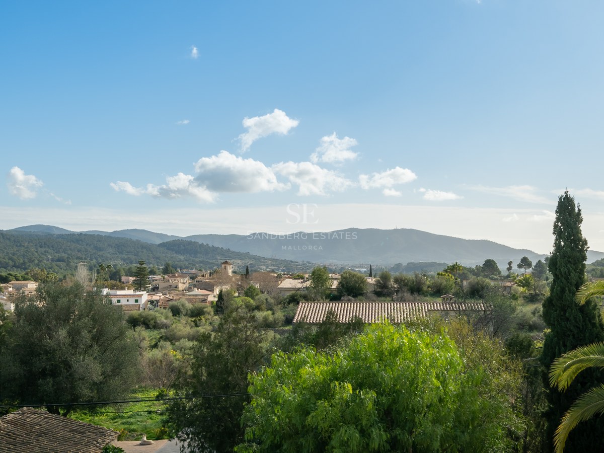 Vista elevada de un pueblo mediterráneo con tejados de terracota rodeado de vegetación y montañas verdes bajo cielo azul.