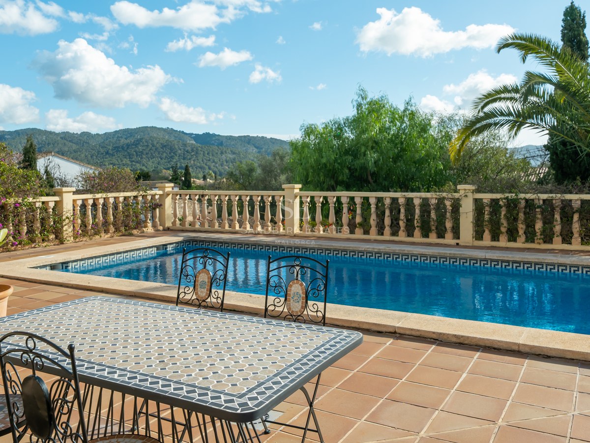 Piscina rectangular y mesa de mosaico en una terraza de terracota con vistas a la montaña y palmeras.