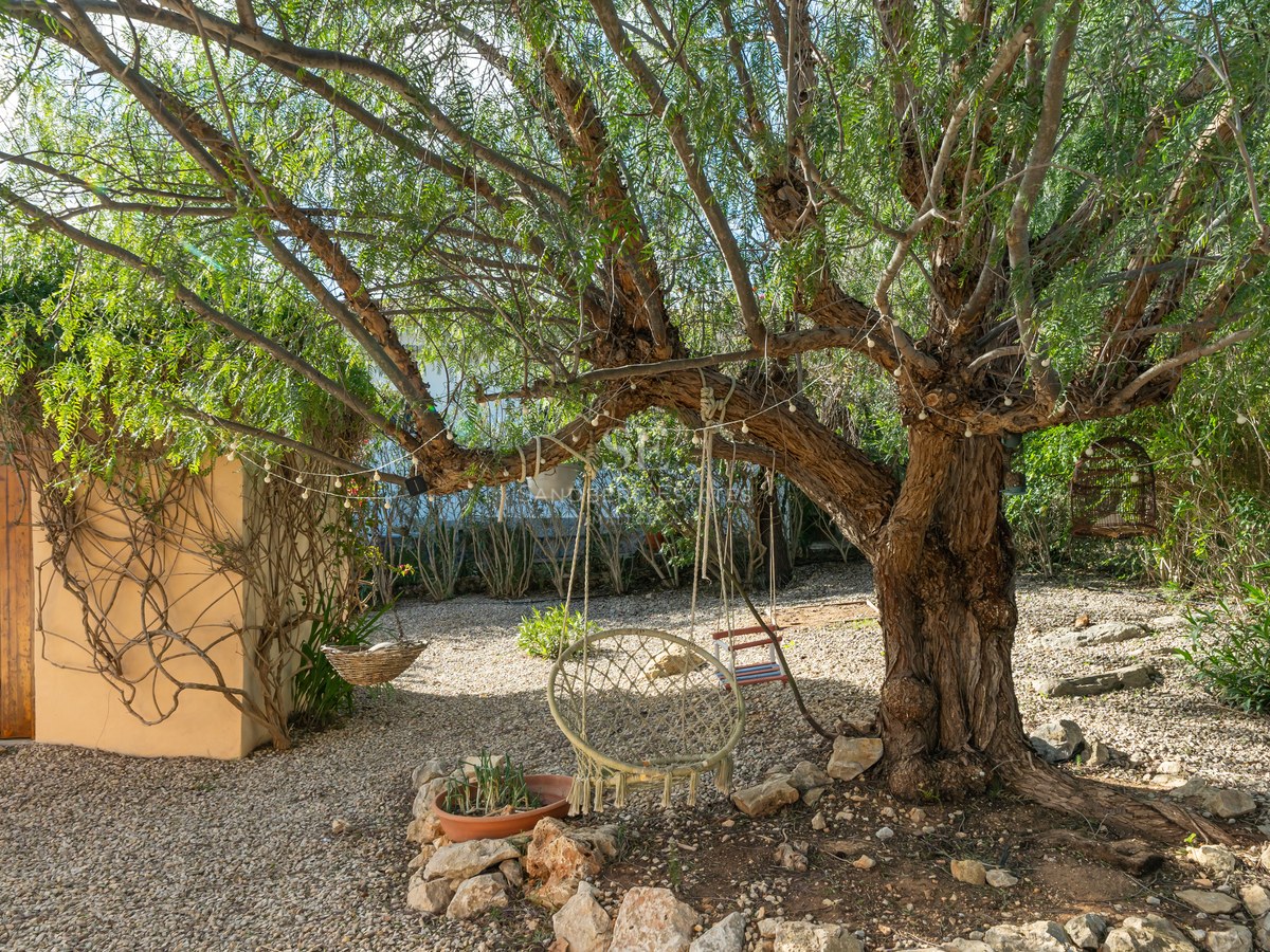 Gran árbol con columpio de macramé y guirnaldas de luces en un jardín de grava junto a una casita de piedra.