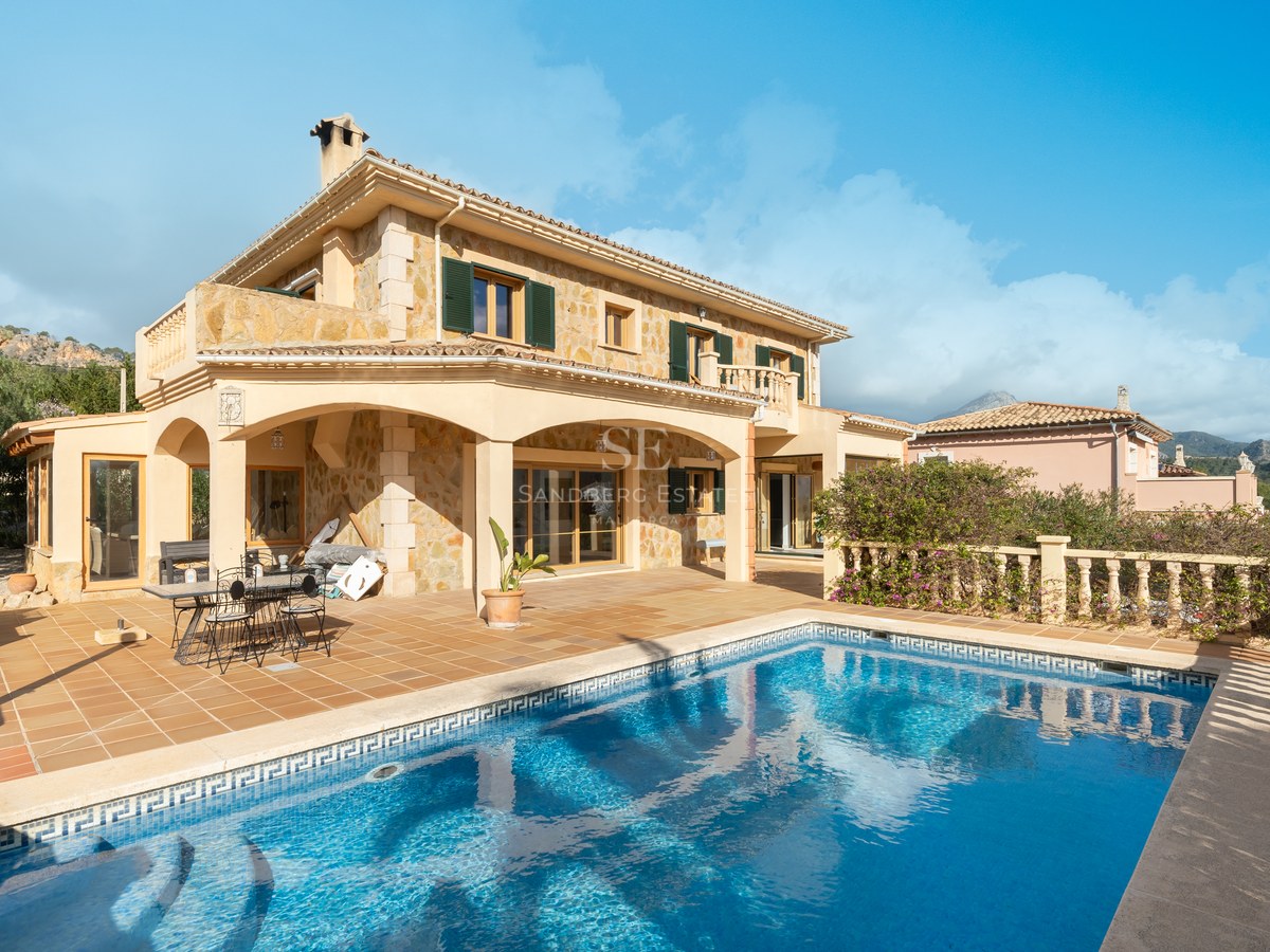 Vista de una villa de piedra de dos plantas con piscina color turquesa y terraza de terracota bajo un cielo azul.
