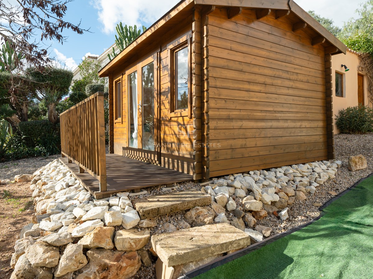 Cabaña de troncos de madera con terraza pequeña en un jardín mediterráneo con muros de piedra natural.