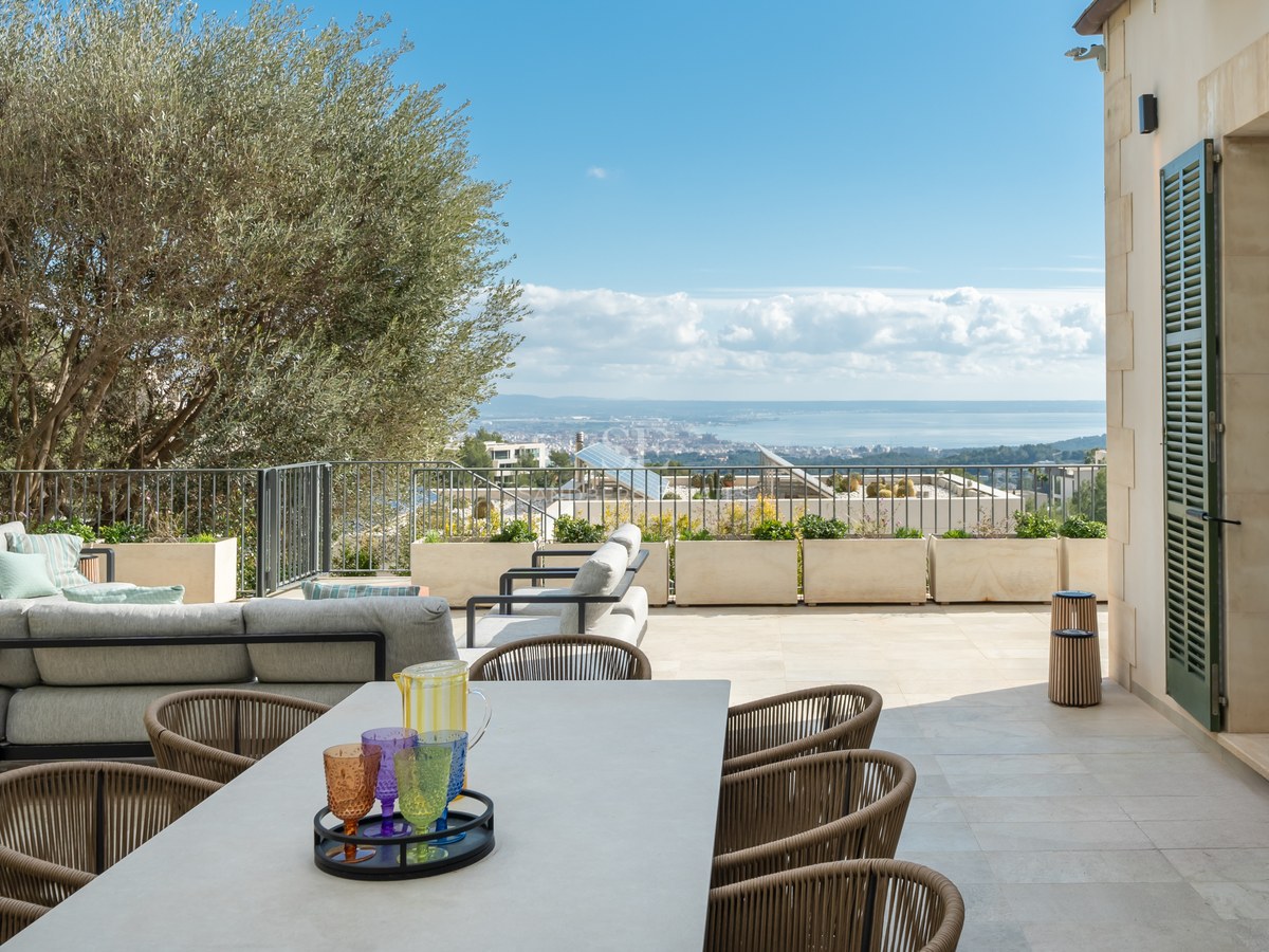 Terraza de piedra con comedor, sofás y olivo con vistas al mar y a la ciudad bajo un cielo despejado.